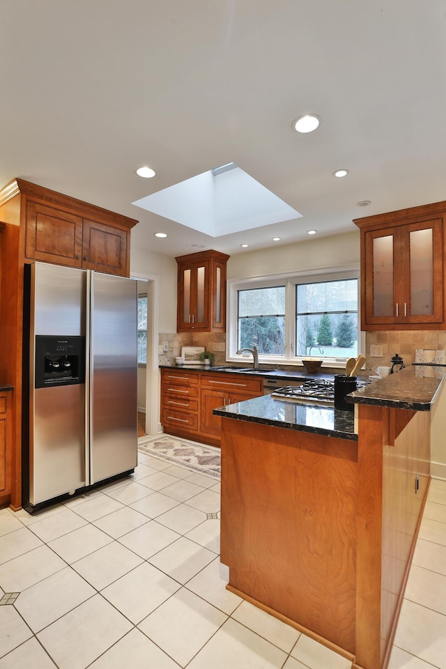 kitchen featuring a skylight, tasteful backsplash, glass insert cabinets, dark stone counters, and stainless steel fridge