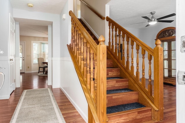 staircase featuring a textured ceiling, ceiling fan, a wainscoted wall, and wood finished floors