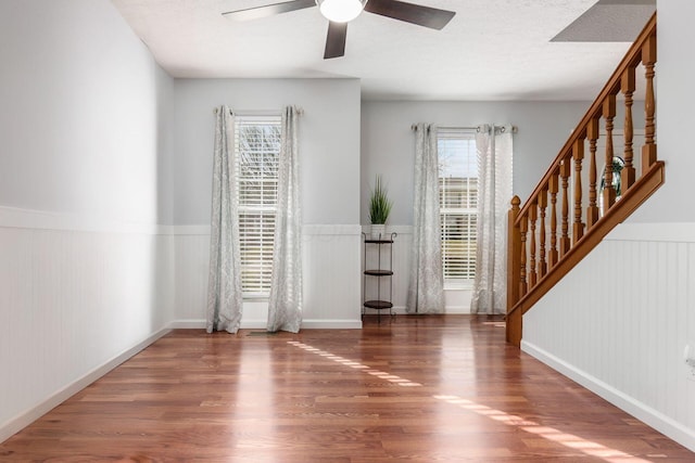 empty room with a textured ceiling, a wainscoted wall, wood finished floors, a ceiling fan, and stairs