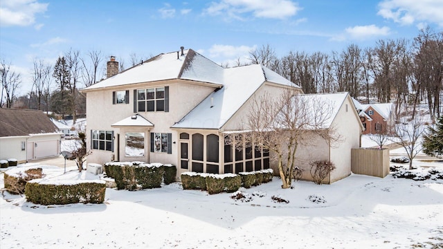 snow covered property with a garage, a sunroom, a chimney, and stucco siding