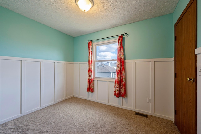 unfurnished room featuring visible vents, a wainscoted wall, a textured ceiling, carpet flooring, and a decorative wall