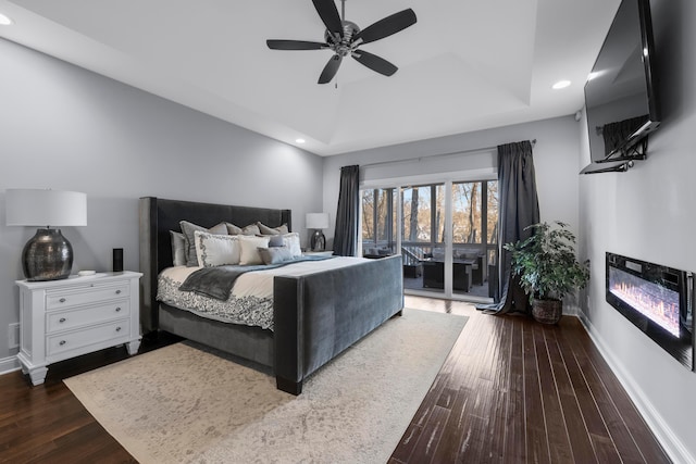 bedroom featuring baseboards, a raised ceiling, a glass covered fireplace, wood finished floors, and recessed lighting