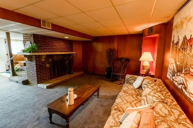 carpeted living room with wood walls, a brick fireplace, and a drop ceiling