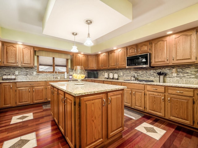 kitchen featuring stainless steel microwave, a center island, brown cabinetry, dark wood finished floors, and decorative light fixtures