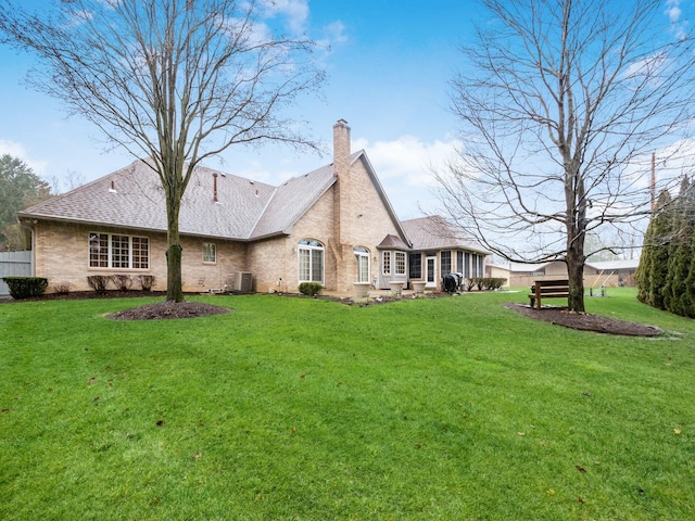 rear view of house featuring central AC unit, brick siding, a shingled roof, a lawn, and a chimney