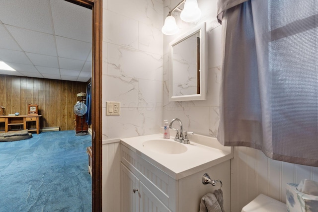 bathroom featuring baseboard heating, vanity, wooden walls, and a drop ceiling