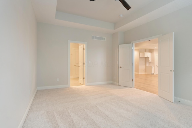 unfurnished bedroom with ceiling fan, ensuite bath, a tray ceiling, and light carpet