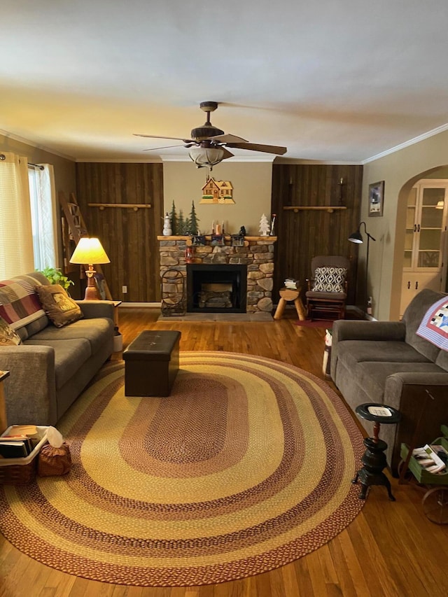 living room featuring crown molding, ceiling fan, wood-type flooring, a stone fireplace, and wood walls