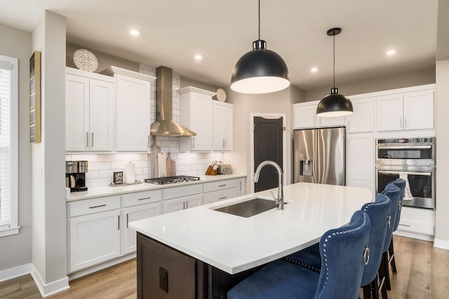 kitchen featuring wall chimney exhaust hood, sink, hanging light fixtures, an island with sink, and stainless steel appliances