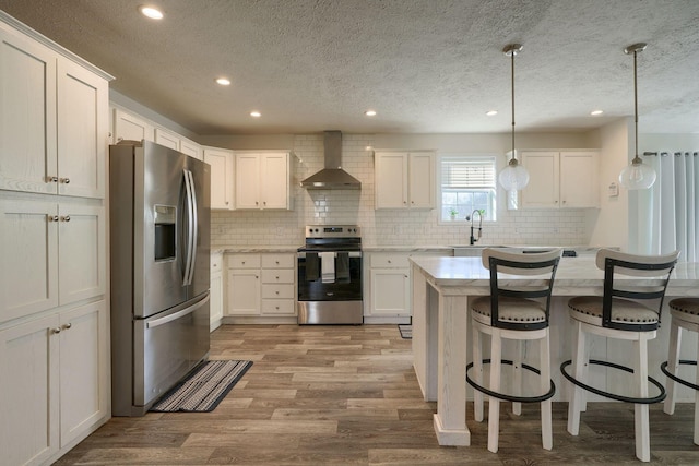 kitchen featuring stainless steel appliances, white cabinetry, hanging light fixtures, and wall chimney range hood