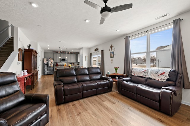 living room with ceiling fan, light wood-style floors, stairs, visible vents, and a textured ceiling