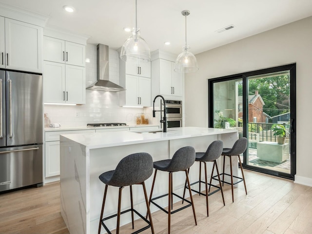 kitchen with sink, a kitchen island with sink, white cabinetry, stainless steel appliances, and wall chimney exhaust hood