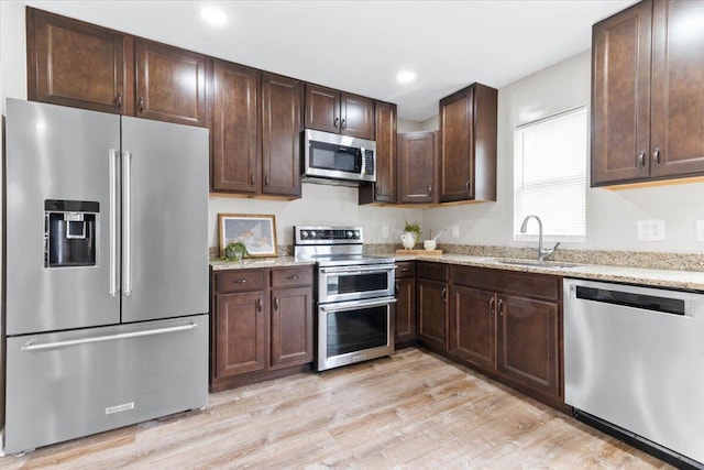 kitchen featuring stainless steel appliances, a sink, dark brown cabinets, light wood-type flooring, and light stone countertops