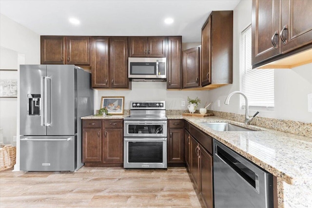 kitchen featuring dark brown cabinetry, light stone countertops, stainless steel appliances, light wood-style floors, and a sink