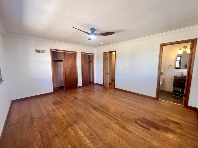 unfurnished bedroom featuring two closets, wood-type flooring, ceiling fan, crown molding, and ensuite bath