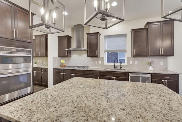 kitchen with wall chimney range hood, sink, stainless steel appliances, light stone counters, and decorative light fixtures