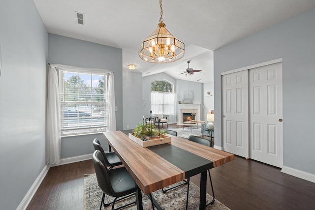 dining room with ceiling fan with notable chandelier, dark wood-type flooring, a fireplace, visible vents, and vaulted ceiling