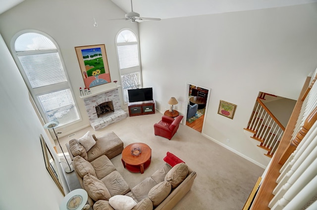 carpeted living area with a fireplace with raised hearth, baseboards, ceiling fan, stairway, and high vaulted ceiling