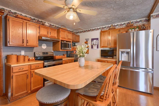 kitchen featuring light wood finished floors, brown cabinets, light countertops, under cabinet range hood, and appliances with stainless steel finishes