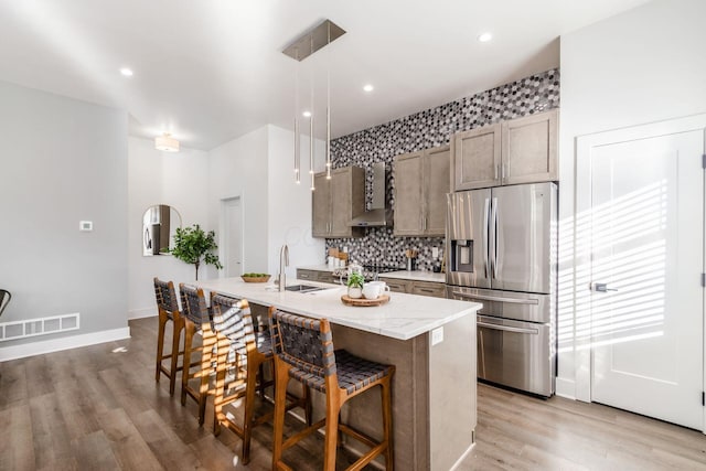 kitchen with a kitchen island with sink, a breakfast bar, decorative light fixtures, stainless steel fridge, and wall chimney range hood