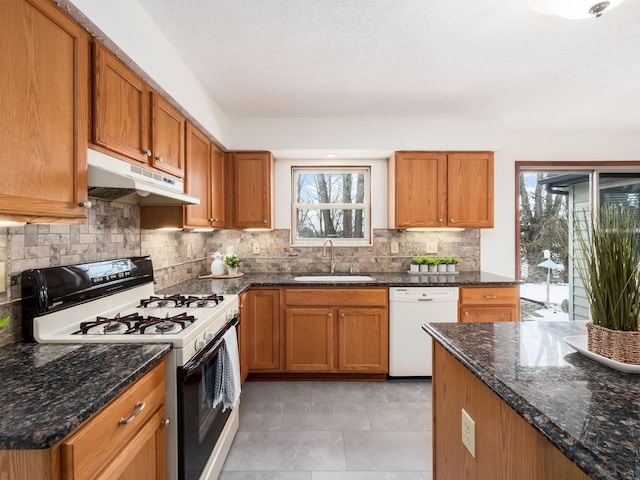 kitchen featuring brown cabinetry, a sink, dark stone counters, white appliances, and under cabinet range hood