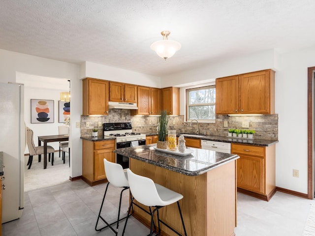 kitchen featuring under cabinet range hood, white appliances, a sink, a center island, and brown cabinetry