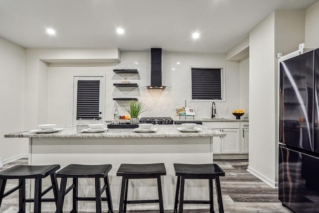 kitchen featuring a center island with sink, black refrigerator, light stone counters, white cabinets, and wall chimney range hood