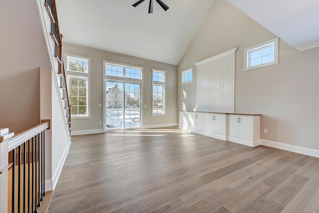 unfurnished living room with ceiling fan, high vaulted ceiling, light wood-style flooring, baseboards, and stairs