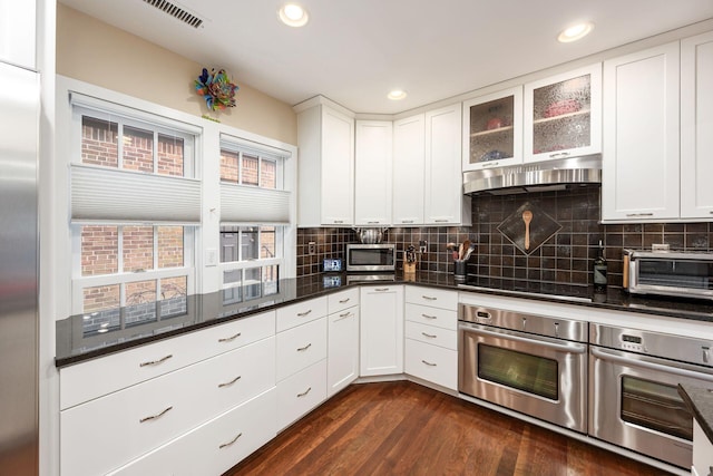 kitchen with under cabinet range hood, dark wood-style flooring, white cabinets, dark stone countertops, and glass insert cabinets