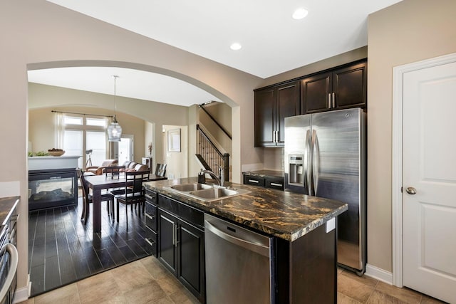 kitchen with dark stone countertops, hanging light fixtures, a kitchen island with sink, stainless steel appliances, and a sink