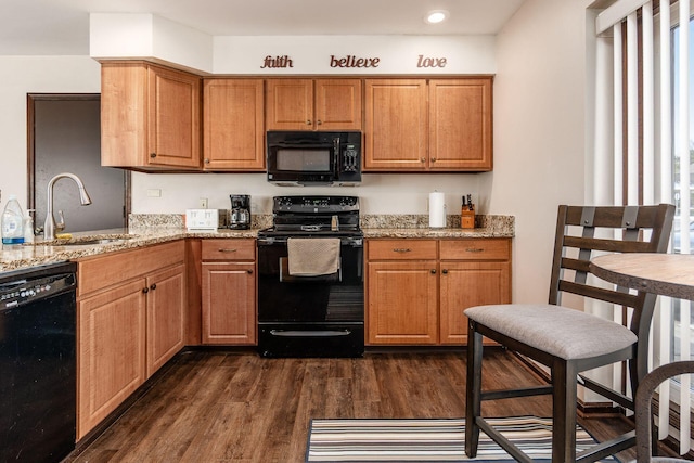 kitchen with brown cabinetry, dark wood-style flooring, light stone countertops, black appliances, and a sink