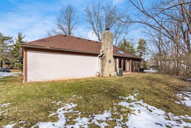 snow covered property featuring a shingled roof, central AC unit, a lawn, and stucco siding