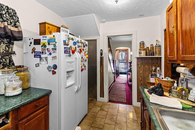 kitchen featuring arched walkways, white refrigerator with ice dispenser, dark countertops, brown cabinetry, and a textured ceiling