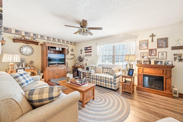 living room with light wood-type flooring, a glass covered fireplace, ceiling fan, and a textured ceiling