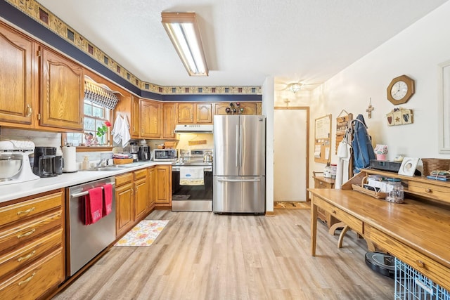 kitchen with stainless steel appliances, light wood-style flooring, brown cabinetry, a sink, and under cabinet range hood