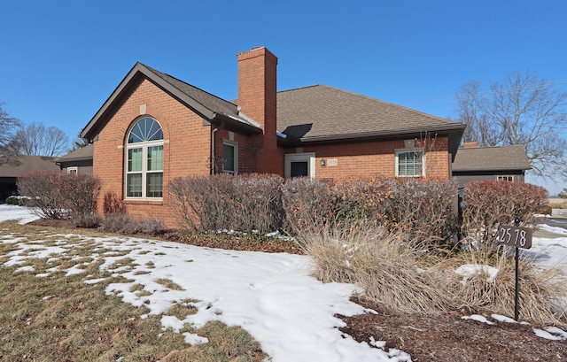 snow covered property with roof with shingles, a chimney, and brick siding