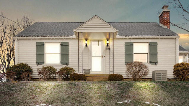 bungalow-style home featuring entry steps, roof with shingles, a chimney, and central air condition unit