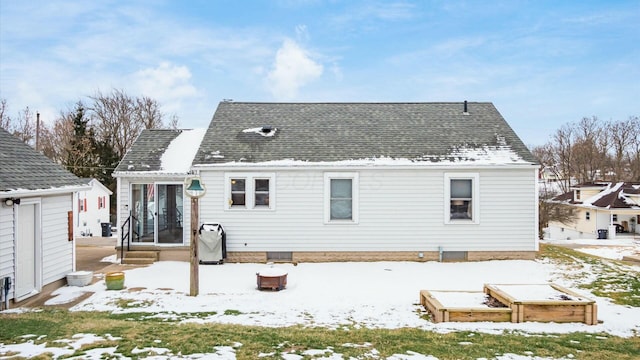 snow covered back of property with roof with shingles and a vegetable garden