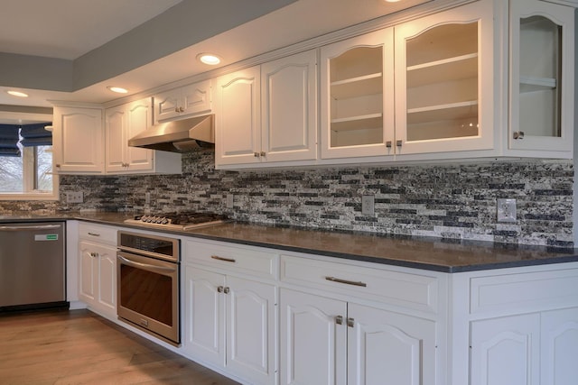 kitchen featuring stainless steel appliances, dark countertops, backsplash, white cabinetry, and under cabinet range hood