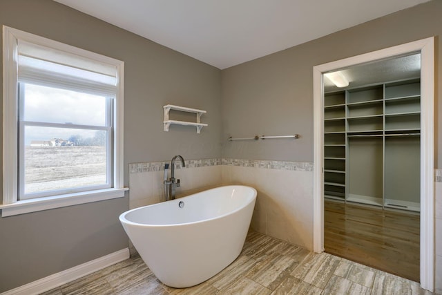 bathroom with a wainscoted wall, wood finished floors, tile walls, a freestanding bath, and a walk in closet