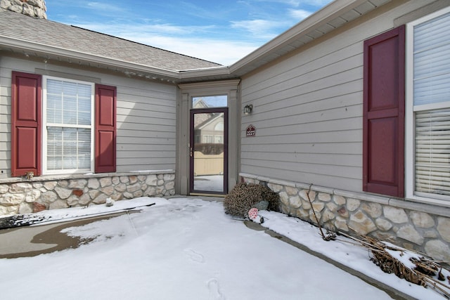 snow covered property entrance with stone siding and roof with shingles