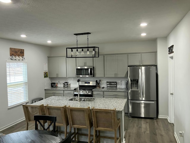 kitchen featuring a kitchen island with sink, gray cabinetry, appliances with stainless steel finishes, light stone countertops, and decorative light fixtures