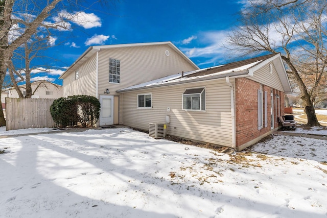 snow covered rear of property featuring central AC, brick siding, and fence