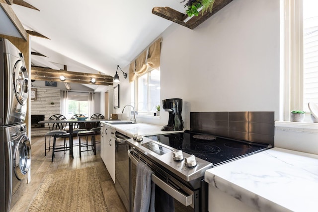 kitchen with stacked washer and clothes dryer, vaulted ceiling with beams, light wood finished floors, appliances with stainless steel finishes, and a sink
