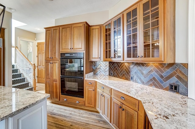 kitchen with glass insert cabinets, brown cabinetry, dobule oven black, and light stone counters