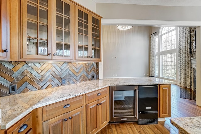 kitchen with light wood-style flooring, beverage cooler, brown cabinets, light stone countertops, and glass insert cabinets