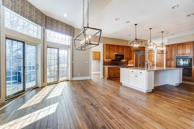 kitchen featuring decorative light fixtures, light wood-style flooring, brown cabinetry, an island with sink, and black appliances