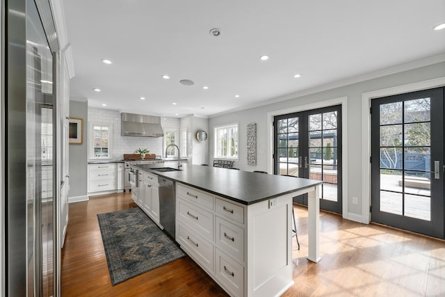 kitchen with an island with sink, dark countertops, wall chimney exhaust hood, french doors, and white cabinetry