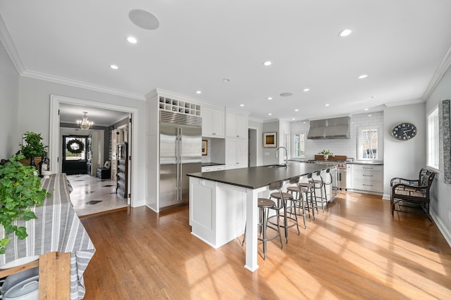 kitchen with built in refrigerator, a sink, wall chimney range hood, dark countertops, and a large island with sink