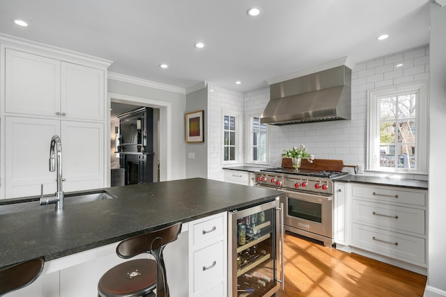 kitchen with designer range, wall chimney exhaust hood, white cabinets, beverage cooler, and a kitchen bar